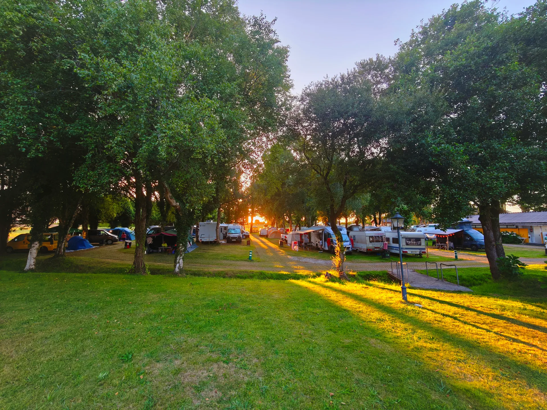 Área de camping con parcelas ocupadas entre árboles, al atardecer.
