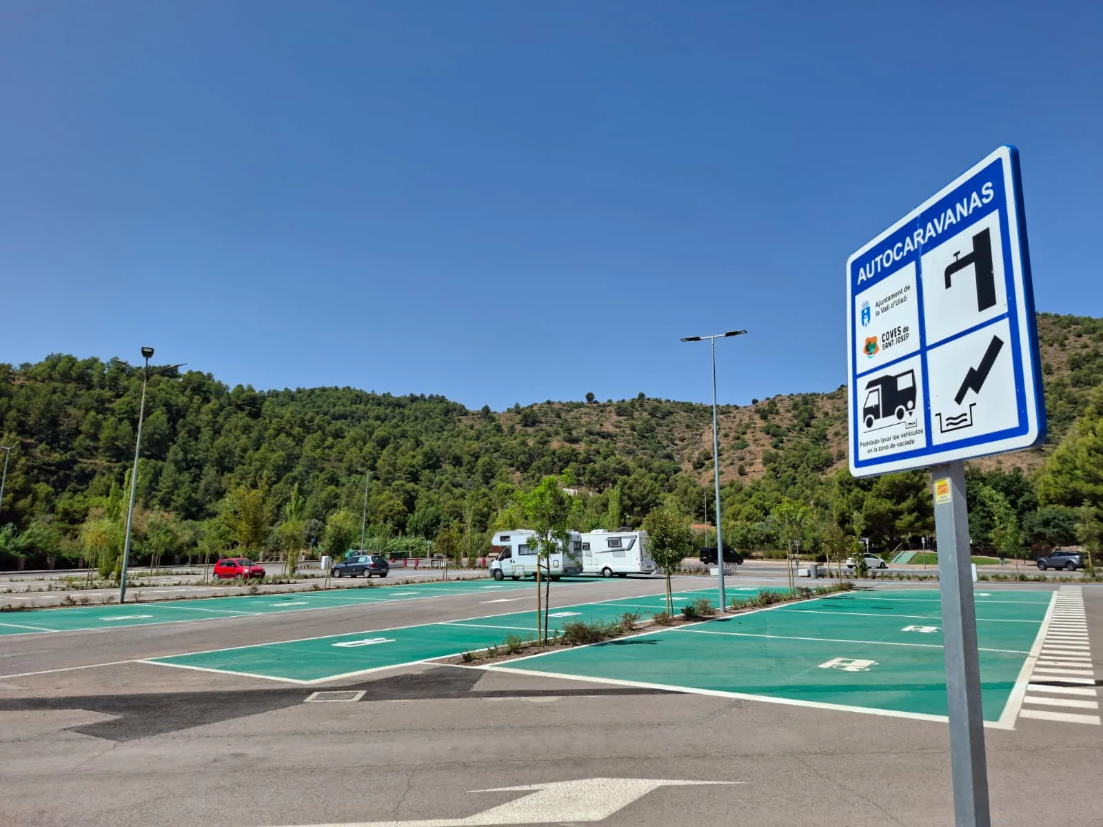 Área de autocaravanas con plazas señalizadas y cartel informativo, junto a una ladera arbolada.