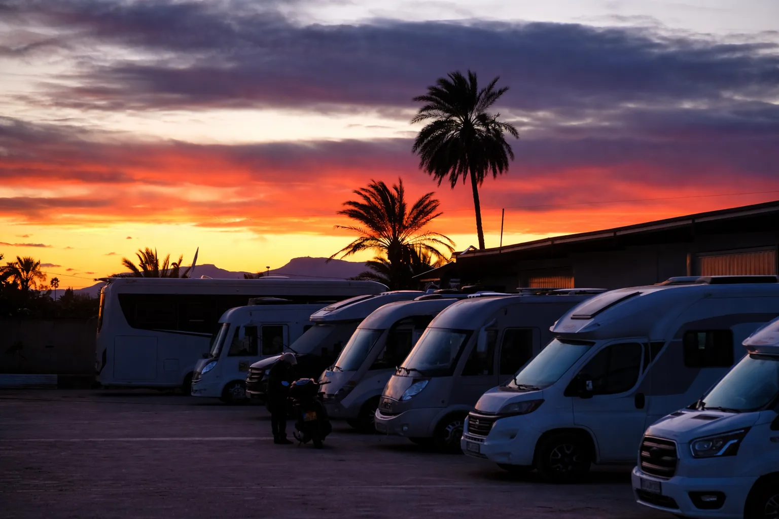 Área de autocaravanas al atardecer, con varias campers estacionadas y palmeras al fondo.