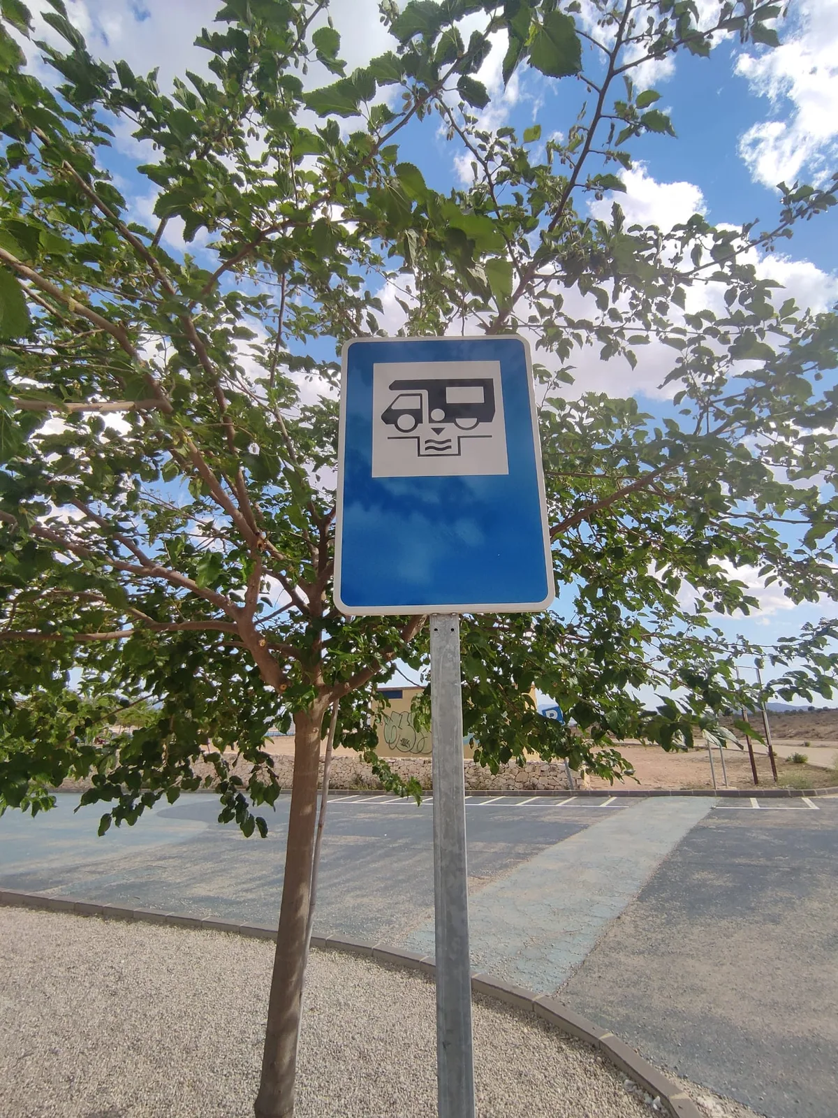 Señal del área de autocaravanas de Yecla junto a un árbol, con el recinto y el paisaje al fondo