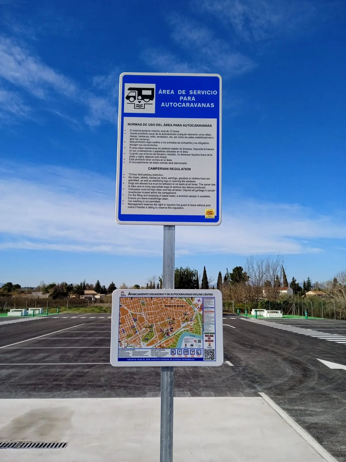 Cartel informativo del área de autocaravanas con vista parcial del recinto y plazas de aparcamiento.